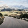 A winding asphalt road curves through a hilly landscape with rocky mountains in the background under a cloudy sky, bordered by stone guardrail posts. Spain freight