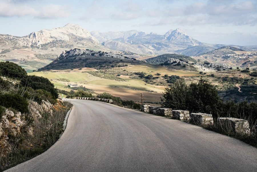 A winding asphalt road curves through a hilly landscape with rocky mountains in the background under a cloudy sky, bordered by stone guardrail posts. Spain freight