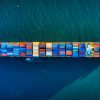 Aerial view of a large container ship sailing in deep blue water, carrying multicolored containers. The scene conveys a sense of scale and global trade.