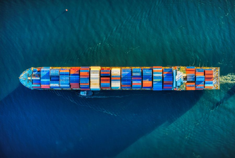 Aerial view of a large container ship sailing in deep blue water, carrying multicolored containers. The scene conveys a sense of scale and global trade.